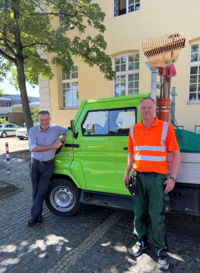 Ein grünes Auto mit Ladefläche ist im mittleren Vordergrund zu sehen. Im Hintergrund ist das gelbe Gebäude des Rathauses. Links steht der Bürgermeister Frank Gellen in einem hell blauen Hemd und lächelt in die Kamera. Rechts im Bild steht der neue Ortspfleger in einer grünen Hose und einer neon orangen Hose.