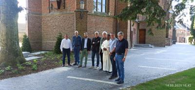Im Hintergrund ist die Brachter Kirche und der Innenhof zu sehen. Die Kirche ist braun und besteht aus Backsteinen. Links im Bild ist ein dicker Baumstamm. Mittig entlang führt ein grau gepflasterter Weg. Acht Herren in unterschiedlicher Kleidung, darunter auch Bürgermeister Frank Gellen, stehen seitlich in einer Reihe und lächeln in die Kamera.