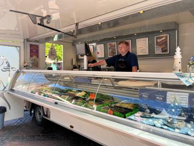 Eine Theke voll mit Fisch und Meeresfrüchten ist zu sehen. Dahinter steht der Fischverkäufer. Der Wagen hat eine weiße Farbe.