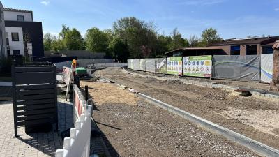 Die Baustelle an der Borner Straße ist abgebildet. Zentral im Bild ist ein Straßenverlauf zu erkennen, bei dem der äußere Rand der Straße mit Steinen eingegrenzt ist. Der Straße ist noch nicht asphaltiert und man sieht Erde. Rechts im Bild steht ein braunes Haus hinter Bauabsperrungen. Links im Bild ist ein asphaltiertes Grundstück mit einem schwarz und weißem Haus zu erkennen sowie eine Mülltonnen-Box. Der Himmel ist blau und im Hintergrund sind grüne Bäume.