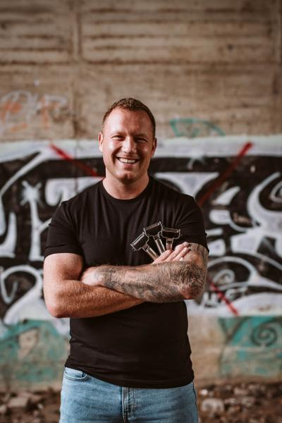 Ein Mann mit schwarzem T-Shirt und einer hellblauen Jeans verschränkt die Arme und lächelt. Im Hintergrund ist eine hell braune Wand die mit einem schwarz und weißem Graffiti aufgesprüht.