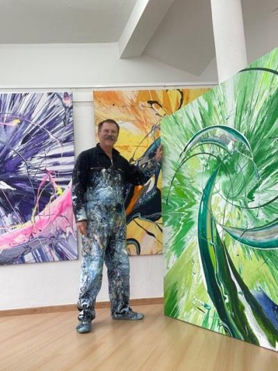 Älterer Mann steht mittig im Bild auf hellen Holzboden mit dunklen Harren, Schnäuzer und Brille trägt dunkle Jacke, Hose und Schuhe, die mit hellen Farben besprenkelt ist, mit linken Hand hält er ein Bild, Bild genauso groß wie Mann und mit grünen Farbtönen, im Hintergrund Ausschnitte von zwei ähnlichen Bildern mit Lila- und Gelbtönen, Bilder hängen an weißer Wand