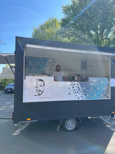 Ein Mann steht in einem schwarzen Anhänger vor der Theke und guckt in die Kamera. Davor hängt ein Schild mit einem Kopf als Symbol und dem Namen Lachma. Rechts befindet sich ein Muster in blau. Im unteren Vordergrund ist ein Kopfsteinpflaster zu sehen und im Hintergrund stehen Bäume und der Himmel ist blau.