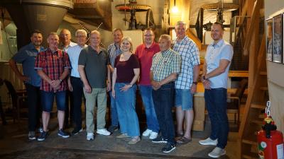 Elf Personen stehen als Gruppe vor einem hölzernen Mahlwerk im Inneren der Brachter Mühle. Im Hintergrund sieht man eine Treppe, Kessel, Trichter und auch ein Mühlenrad und -stein. 
