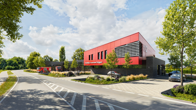 Ein Feuerwehrhaus ist zu sehen. Das Bauwerk hat ein Obergeschoss in rot und ein Untergeschoss in einem anthrazit-Ton. Im Vordergrund ist eine Straße zu sehen und ein zahlreiche Pflanzen und Grünflächen und einige Autos und ein Feuerwehrauto links. Außerdem befinden sich links im Bild Garagenhallen. Auf dem Gebäude steht Freiwillige Feuerwehr Brüggen und die Nummer 112.