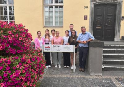 Im Hintergrund sind die gelben Fassaden des Rathauses in Brüggen zu sehen und Fenster sowie auch der Eingang mit den braunen großen Holztüren und die Treppe. Links im Bild steht ein großer Blumenkübel mit pinken Blumen. Mittig im Bild sind fünf junge Damen zu sehen. In der hinteren Reihe sind Sachgebietsleitung Annette Becker, Fachbereichsleitung Sascha Achten und Bürgermeister Frank Gellen abgebildet.