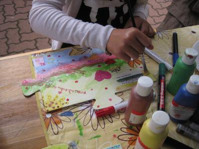 Ein Mädchen malt auf einem Blatt Papier. Zu sehen sind viele verschiedene Buntstifte, Filzstifte, Pinsel und Farben. Im Hintergrund ist ein Holztisch und ein Kopfsteinpflaster zu sehen. Außerdem ist im Vordergrund ein Puzzle Teil zu sehen, dass bereits mit vielen bunten Farben angemalt wurde.