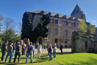 Zu sehen ist eine Burg im Hintergrund mit braunen Backsteinen und einen Burgturm der dunkelgrau ist. Rechts im Bild stehen vor der Burg einige Burgruinen und Pflanzen. Im Vordergrund ist ein grüner Rasen zu sehen, auf dem eine Gruppe Menschen versammelt steht und zuhört. Der Himmel ist blau und wolkenlos.