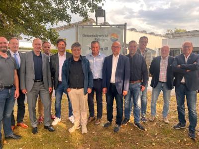Vor dem Gebäude stehen 12 Herren, die alle zusammen in die Kamera lächeln. Im Hintergrund ist das weiße Gebäude der Gemeindewerke Brüggen zu sehen. Im Vordergrund ist ein grüner Rasenabschnitt zu sehen. Außerdem befindet sich links im Bild ein grüner Baum. Der Himmel ist leicht gräulich und bewölkt.