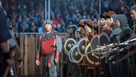Ritter mit rot-grauer Kleidung links von einer Reihe von weiteren Ritten mit Speeren und Schildern vor Zuschauern auf einer Tribüne verkleidet mit Holzlatten