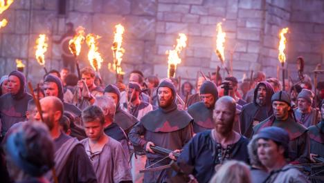 Mittelalterlich gekleidete Menschenmenge mit Fackeln dazwischen Reihe von Rittern mit Armbrüsten vor Steinmauer