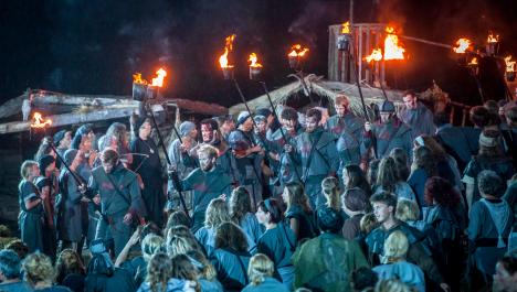 Mittelalterlich gekleidete Menschenmenge im unteren Teil des Bildes (von hinten) schaut auf Ritter mit Fackeln, dahinter weitere mittelalterlich gekleidete Menschen vor Holzhütten bei Nacht