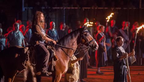 Reiter auf braunem Pferd neben einzelnen Menschen mit Fackeln vor mittelalterlicher Menschenmenge bei Nacht mit Fackellicht