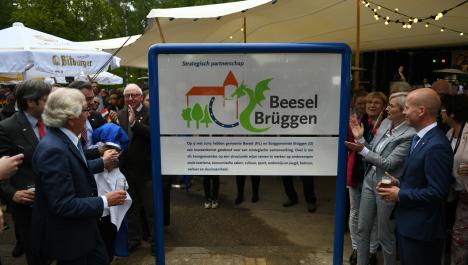 Weißes Schild befestigt an blauer Metallrahmen mit Logo zur Städtepartnerschaft und schwarzem Schriftzug daneben "Beesel Brüggen" darunter Informationstext, links und rechts förmlich gekleidete und feiernden Menschen, im Hintergrund links weißer Sonnenschirm mit goldener Aufschrift "Bitburger" und rechts Zelt mit hellem Segel und Lichterketten, dahinter grüner Baumbewuchs