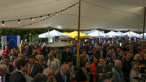 Feiernde Menschenmenge unter großem Zelt mit weißer Decke und Holzpfahl als Stütze in der Mitte, Stehtische mit kleiner Flagge von Deutschland und den Niederlanden sowie Gasheizstrahler, im Vordergrund, im Hintergrund weiße und gelbe Pavillons und Schirmen, 