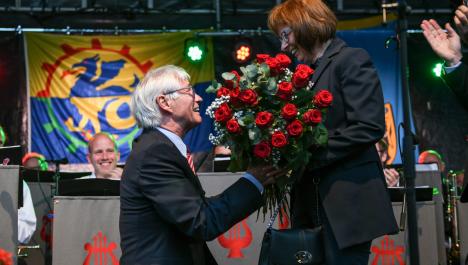 Älterer Mann im Anzug auf Knien links übergibt Frau im schwarzen Hosenanzug mit schwarzer Handtasche einen Strauß mit Rosen auf Bühne vor Musikern, im Hintergrund buntes Bühnenbild