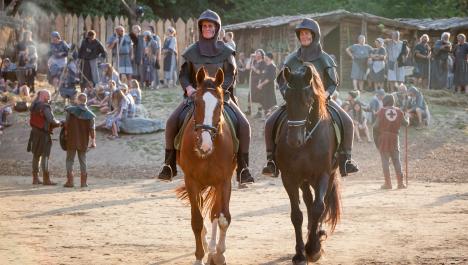 Zwei Ritter auf zwei Pferden, links braun und rechts schwarz, auf erdigem Boden, dahinter mittelalterlich gekleidete Menschen in einem dorf mit Holzhütten rechts und Feuerstelle links, im Hintergrund Zaun bestehende aus Holzlatten, dahinter dichter Baumbewuchs