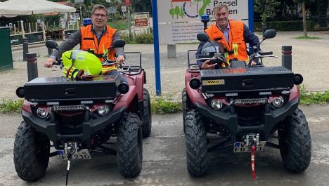 Zwei Männer auf je einem Feuerwehr-Quad mit orangen Warnwesten, dahinter Schild zur Städtepartnerschaft mit Logo und Schriftzug "Beesel-Brüggen" und Informationstext, Parkplatz, Gastronomie und dichtem Baumbewuchs 