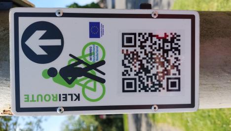 Weißes Schild an Holzpfahl mit QR-Code unten, schwarze Figur mit Wanderstock und dahinter grüne Figur auf grünem Fahrrad, oben recht weißer Pfeil im schwarzem Kreis geradeaus oben, links seitlicher grün, schwarzer Schriftzug "Kleiroute"