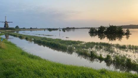 Panoramablick auf mit Wasser überschwemmtes Feld, teilweise Wiesen- und Strauchbewuchs im Wasser, im Hintergrund Mühle links und Himmel beim Sonnenuntergang