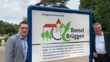 Schild zwischen blauen Rahmen mit Überschrift in blau "Strategische Partnerschaft", darunter Logo Beesel-Brüggen (Umriss Burg orange, unter linker Teil der Burg drei grüne Bäume, rechts davon blauer Halbkreis mit gelben Sternen als Zeichen der Europäischen Union, rechts von der Burg grüner Drache, rechts Schriftzug "Beesel Brüggen") und Informationstext darunter, links und rechts davon je ein Mann im Anzug , im Hintergrund gepflasterter Platz und dichter, grüner Baumbewuchs 