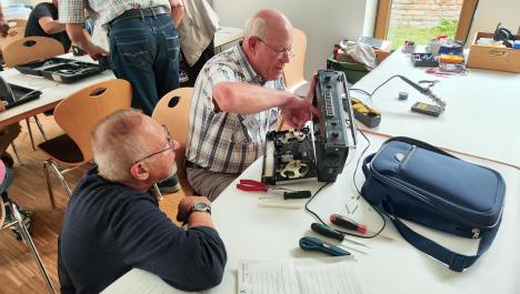 Mittig: Älterer Mann mit Brille und kariertem Hemd sitzt an weißem Holztisch und schraubt an elektronischen Gerät, rechts von ihm Mann mit Brille, Armbanduhr, und Pullover schaut zu, auf Tisch diverse Werkzeuge und ein blauer Rucksack, im Hintergrund links stehen und sitzen Personen an weißem Holztisch und auf Holzstühlen mit Metallbeinen und rechts Holzkiste und Werkzeug auf Tisch vor Fenster mit Holzrahmen mit Blick auf Bachsteinwand und Wiese