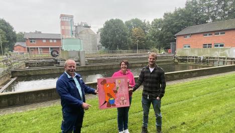 Drei Personen nebeneinander auf grüner Wiese halten rosa Schild, darauf orange Maus mit Pinsel, Farbeimer und Leinwand, Mann links mit blauer Arbeitsjacke und -hose, Arbeitsschuhe, hellblau-weiß gestreiftes T-Shirt, Frau in der Mitte mit pinken Pullover, blauer Jeans , weißen Turnschuhen, Mann rechts mit dunkel karierten Hemd, darunter weißes T-Shirt, blaue Jeans, Arbeitsschuhe, im Hintergrund großes Becken mit Wasser, dahinter Kläranlage mit roten Gebäuden, dichter Baumbewuchs, bei grau bewölktem Himmel