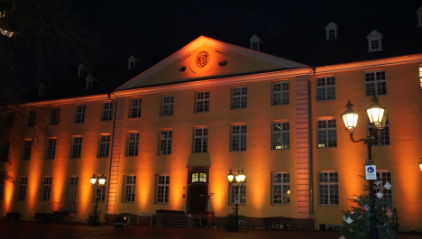 Großes Gebäude mit vielen Fenster wird mit orangen Lichtstrahlern beleuchtet, mittig unten Eingangstür mit Treppenaufgang und oben Wappen von Brüggen, vor Gebäude gepflasterter Platz mit drei leuchtenden Laternen mit je drei Leuchteinheiten, an Laternen Tannenbäume befestigt, Aufnahme bei Nacht