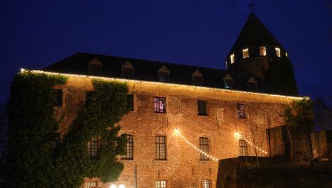 Angestrahlte Burg Brüggen mit Lichterkette an Regenrinne, rechts Licht in Turmfenstern und Efeu links an der Fassade, rechts vor der Burg dunkle Mauer und kleine Ausschnitte von Weihnachtsmarkthütten und runden leuchtenden Straßenlaternen, dunkler Himmel bei Nacht