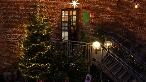 Stahltreppe zur Eingangstür der Burg Brüggen, dinnen Licht an, oben auf der Treppe Person in Weihnachtskleidung, unten vor der Treppe Straßenlaterne mit zwei runden leuchtendenden Glaskugeln und Ausschnitt von Menschenmenge, hinter Menschen und vor Burg großer grüner Tannenbaum geschmückt mit Weihnachtskugeln und Lichterkette, links daneben Holzschlitten zum rein setzen, Burgwand im Hintergrund aus roten Backstein 