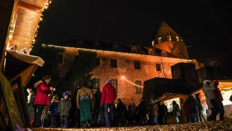 Bildaufnahme vom Boden aus von der angestrahlten Burg Brüggen mit Lichterkette an Regenrinne, rechts Licht in Turmfenstern und Efeu links an der Fassade, rechts vor der Burg dunkle Mauer, links und rechts Weihnachtsmarkthütten aus Holz, zwischen den Hütten Menschen in dicker Winterkleidung, Boden mit braunen Rindenmulch, dunkler Himmel bei Nacht