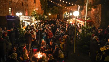 Menschenmenge in dicker Winterkleidung im Burginnenhof, im Vordergrund sitzen Kinder mit Stockbrot im Kreis um Feuerschale, zwischen Menschenmenge leuchtende Straßenlaternen und Tannenbäume, links Stand mit Essen, rechts beleuchtete Weihnachtsmarkthütte aus Holz, links Burg Brüggen mit Licht in Fenster und Stahltreppe zum Eingang, rechts Burgmauer, im Hintergrund Eingangstor zum Innenhof, von links nach rechts Lichterkette gespannt, dunkler Himmel bei Nacht 