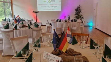 Raum mit Holzboden, Stühlen bezogen mit weißen Hussen an runden gedeckten Tischen mit weißer Tischdecke und deutschen, niederländischen und europäischen Flaggen und Tic-Tacs mit Flaggen beklebt, im Hintergrund steht Beamer auf Standfuß, Präsentation auf Wand projiziert, links und rechts davon farbige Strahler, rechts weiße Wand mit Tür und Lampen, links Fensterfront, oben Holzdecke mit weißer Deko und Lampen