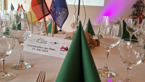Im Vordergrund runder gedeckter Tisch mit weißer Tischdecke und deutschen, niederländischen und europäischen Flaggen und Tic-Tacs mit Flaggen beklebt, Schild zum Arbeitstreffen der Räte 24. Juni 2024 mit Logos, im Hintergrund weitere gedeckte Tische, Beamer steht auf Standfuß, Präsentation auf Wand projiziert, links und rechts davon farbige Strahler, Tür und Lampen, links Fenster, oben links weiße Deko