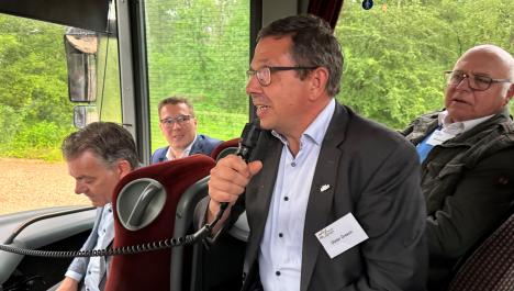 Vier Männer sitzen in Reisebus vorne rechts, im Vordergrund Mann mit Brille, grauen Sacko, weißem Hemd und dunkler Hose hält Mikrofon in der Hand, Hinter den Männern Fenster, vorne links Frontscheibe mit Außenspiegeln, im Hintergrund dichter grüner Baum- und Strauchbewuchs 