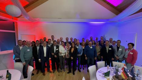 Menschen stehen für Gruppenfoto zusammen auf Holzboden, davor zwei Runde gedeckte Tische mit weißen Stühlen, weißer Tischdecke und deutschen, niederländischen und europäischen Flaggen und Getränke, im Hintergrund Wand beleuchtet mit zwei farbigen Strahlern, rechts weiße Wand mit Tür, links Fensterfront, oben Holzdecke mit weißer Deko