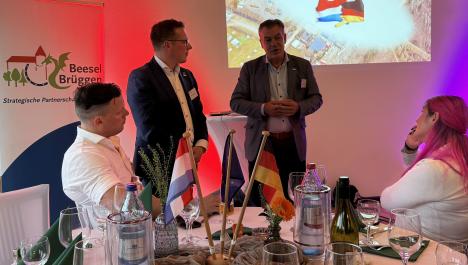 Im Vordergrund sitzen links ein Mann in weißem Hemd und rechts eine Frau in weißem Oberteil mit pinken Haaren auf Stühlen mit weißen Hussen an einem gedeckten Tisch mit weißer Tischdecke und deutschen, niederländischen und europäischen Flaggen, Präsentation auf Wand projiziert, links und rechts davon farbige Strahler, links stehen zwei Männer in Anzügen (Bürgermeister von Beesel und Brüggen), links Aufsteller in weiß-blau mit Logo und Schriftzug zur Städtepartnerschaft von Beesel und Brüggen
