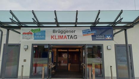 Eingang Burggemeindehalle Brüggen: Eingangstür bestehend aus zwei Flügeltüren aus Glas stehen offen, Blick in die Halle auf geschlossene Glastür, davor Teppichleiste und silberne Standsäulen, links und rechts von der Tür Lampen und zwei bodentiefe Fenster mit Milchglas, darüber Vordach aus Glas und Metallgerüst, daran hängt Banner zum Klimatag, links auf Banner Logo Klimatag, mittig Schriftzug Klimatag und rechts Volksbank Logo, oben bewölkter Himmel