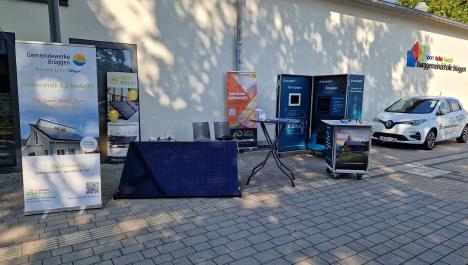 Stand vor Gebäude mit weißer Fassade auf grau gepflasterten Boden, links vorne Werbeaufsteller der Gemeindewerke Brüggen, daneben rechts aufgestelltes Solarpanel, rechts daneben Stehtisch mit Infomaterial, dahinter Werbeaufsteller Hepa Solar und zwei grau Stühle, rechts davon zwei Rollwagen zu Solarwatt, dahinter Werbeaufsteller Elektro Dings, ganz rechts weißes Auto Renault Zoe, darüber an der Wand Logo der Burggemeindehalle, im Hintergrund oben rechts blauer Himmel und grüne Baumkrone