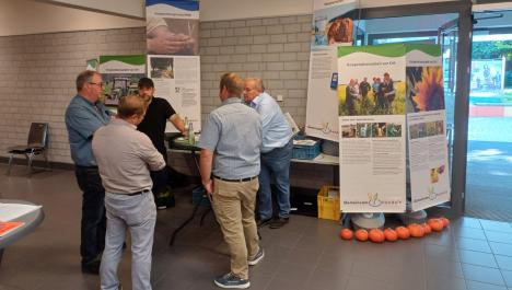 Aussteller: links und rechts je drei Werbeaufsteller, rechts vor Aufstellern 9 orange Kürbisse, dazwischen Tisch mit Kisten, davor Stehtisch mit Getränken, darum stehen vier Männer mit Hemden, einer mit Cap und schwarzem T-Shirt, in langen Hosen, rechts Eingangstür Burggemeindehalle, graue Steinwand, weiße Wand, grauer Fliesenboden