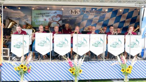 Orchester mit Instrumenten auf Bühne, vor sitzenden Personen 7 weiße Wimpel mit Logo Noten Krakers, Bühne bayrisch dekoriert, Rückwand mit weiß-blau karierter Abdeckung, Werbung für Trachtenmode Niederkrüchten, links Bildschirm mit „Städtepartnerschaft Beesel-Brüggen Grenzenloos feiern“, vorne weiß-blau karierte Abdeckung, vor Bühne drei Vasen mit Blumensträußen, u.a. Sonnenblumen auf Wiese