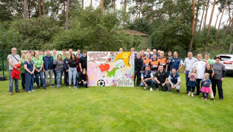 Gruppe von Menschen stehen links und rechts von bunt bemalter Leinwand auf grüner Wiese, oben links auf Leinwand steht Wort „Brüggen“ und unten rechts „Beesel“, im Hintergrund dichter grüner Baum- und Strauchbewuchs, dazwischen Ausschnitt von grünem Haus, rechts Ausschnitt vom weißen Auto