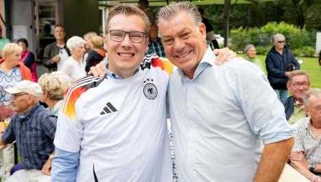 Bürgermeister Bob Vostermans aus Beesel mit Brille, weißen Deutschlandtrikot über hellblauen Hemd links und Bürgermeister Frank Gellen mit weißem Hemd rechts haben Arme umeinander gelegt, im Hintergrund sitzen und stehen Menschen, Bierpavillon grüne Wiese und grüner Strauchbewuchs
