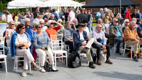 Menschen sitzen auf weißen Stühle auf grauer Steinterasse, einzelne Stühle frei, im Hintergrund stehen Menschen an dekorierten Stehtischen mit weißen Hussen, links weiße Schirme, rechts Pavillion
