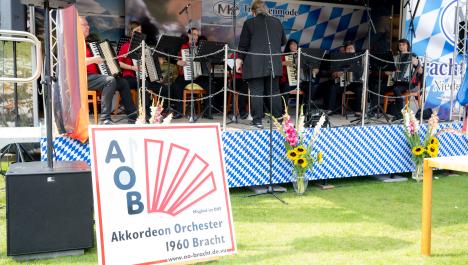 Orchester mit Akkordeons auf Stühlen mit Notensätndern auf Bühne, Bühne bayrisch dekoriert, Rückwand mit weiß-blau karierter Abdeckung, Werbung für Trachtenmode Niederkrüchten, links Bildschirm mit „Städtepartnerschaft Beesel-Brüggen Grenzenloos feiern“, vorne weiß-blau karierte Abdeckung, links Deutschlandfahne, rechts niederländische Fahne, vor Bühne zwei Vasen auf grüner Wiese mit Blumensträußen, u.a. Sonnenblumen, links schwarzer Lautsprecher, rechts daneben Schild mit Logo Akkordeon Orchester Bracht