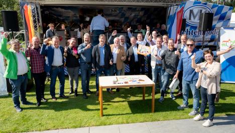 Gruppe von Menschen haben einen Arm in der Luft oder zeigen Daumen nach oben und stehen vor bayrisch geschmückter Bühne, links Deutschlandfahne, rechts niederländische Fahnevor ihnen Tisch mit Puzzle steht auf grüner Wiese, davor graue Steinterasse, im Hintergrund Orchester auf Bühne
