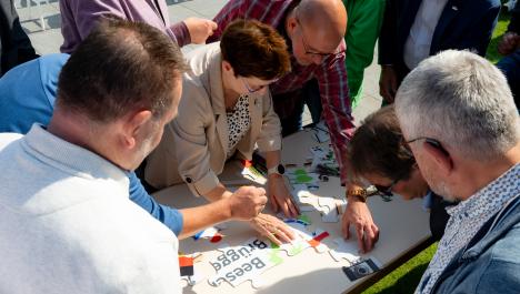 Sicht von oben, Menschen stehen um Tisch und puzzlen gemeinsam ein großen Puzzle, einzelne und teils schon zusammenhänge Puzzleteile liegen auf Tisch, Tisch steht auf grüner Wiese