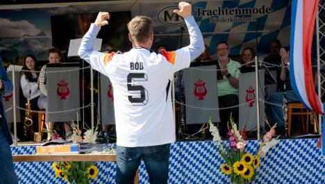 Bürgermeister Bob Vostermanns (Beesel) steht mit Rücken zur Kamera und zeigt mit Daumen auf seinen Rücken, er trägt weißen Deutschlandtrikot mit „Bob“ und Nr. 5 über hellblauem Langarmhemd und blauer Jeans, im Hintergrund Bühne bayrisch dekoriert, Rückwand mit weiß-blau karierter Abdeckung, Werbung für Trachtenmode Niederkrüchten, links Bildschirm, vorne weiß-blau karierte Abdeckung, rechts niederländische Fahne, vor Bühne zwei Vasen mit Blumensträußen, u.a. Sonnenblumen, davor Holztisch mit Puzzlekarton
