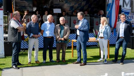 4 Männer vom Verein Stichting Grenzenloos links, rechts Bürgermeister Frank Gellen (Brüggen) und Bob Vostermans (Beesel), dazwischen Frau stehen auf grüner Wiese und grauer Steinterasse, im Hintergrund Orchester auf Bühne, Bühne bayrisch dekoriert, Rückwand mit weiß-blau karierter Abdeckung, links Bildschirm, vorne weiß-blau karierte Abdeckung, links Deutschlandfahne, rechts niederländische Fahne sowie Lautsprecher, vor Bühne 3 Vasen mit Blumensträußen, davor Holztisch auf grüner Wiese