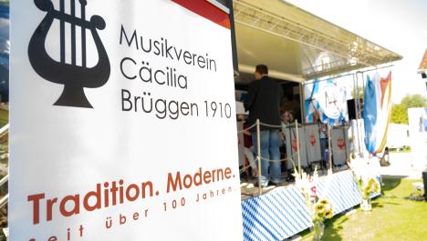 Links im Vordergrund Aufsteller vom Musikverein Cäcilia Brüggen 1970, rechts im Hintergrund Orchester und Dirigent auf Bühne, grau-rote Wimpel vor Stühlen, Bühne bayrisch dekoriert, vorne weiß-blau karierte Abdeckung, rechts niederländische Fahne sowie Lautsprecher, vor Bühne 2 Vasen mit Blumensträußen auf grüner Wiese